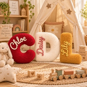May include: Three plush letter-shaped cushions in red, cream, and mustard yellow. The red C features the name Chloe, the cream D features the name Daisy, and the mustard L features the name Lily in raised script lettering on the front surface.