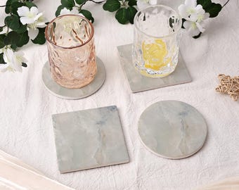 May include: Two textured glass tumblers, one pink and one clear with a yellow base, each resting on a stone coaster. Two additional stone coasters, one square and one circular, feature a marbled grey and white pattern with subtle blue veining.