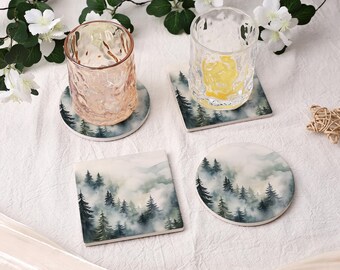 May include: Two clear, textured drinking glasses resting on two round coasters, alongside one square coaster and one round coaster, all featuring a printed design of dark green pine trees set against a backdrop of soft, misty white clouds.