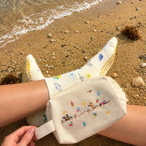May include: A cream-colored canvas cosmetic bag with a white handle, embroidered with figures doing yoga on a beach. The person is wearing cream-colored socks with yellow grips and floral designs. The scene is set on a sandy beach.