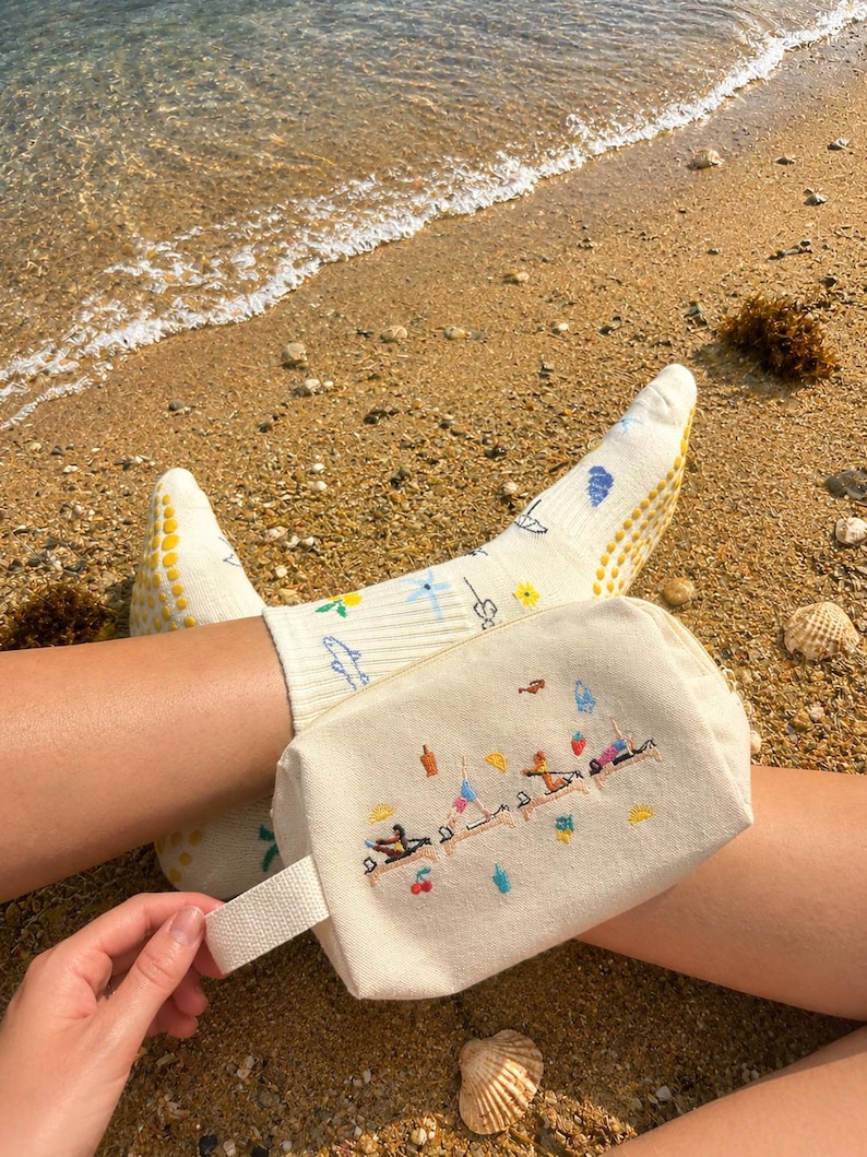 May include: A cream-colored canvas cosmetic bag with a white handle, embroidered with figures doing yoga on a beach. The person is wearing cream-colored socks with yellow grips and floral designs. The scene is set on a sandy beach.