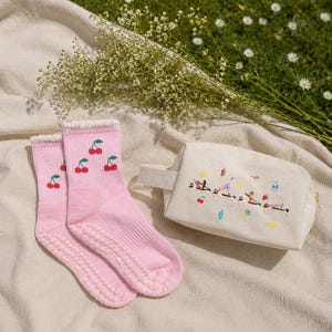 May include: A pair of pink socks with cherry embroidery and grip soles, next to a cream-colored cosmetic bag with embroidered figures and objects. The items are arranged on a beige blanket with a green grass background.