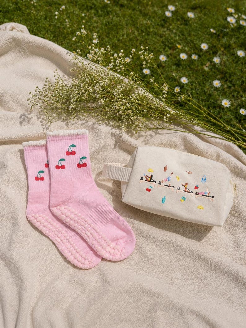 May include: A pair of pink socks with cherry embroidery and grip soles, next to a cream-colored cosmetic bag with embroidered figures and objects. The items are arranged on a beige blanket with a green grass background.