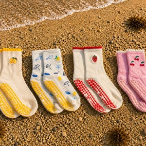 Könnte beinhalten: Vier Paar Socken mit gerippten Bündchen und rutschfesten Noppen an den Sohlen. Das erste Paar ist cremefarben mit gelben Zitronen, das zweite weiß mit blauen und gelben Strandmotiven, das dritte weiß mit einer roten Erdbeere und das vierte rosa mit roten Kirschen.
