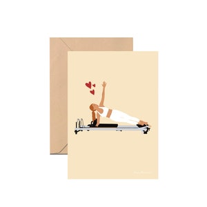 May include: A greeting card featuring a cartoon illustration of a person performing a pilates exercise on a reformer machine. The person is wearing white clothing and there are red hearts floating above them.
