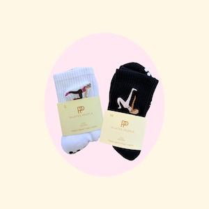 May include: Two pairs of Pilates People socks, one white and one black. The white socks have a pink and black embroidered figure in a yoga pose, and the black socks have a white embroidered figure. Both pairs have a tan label with the brand name.