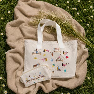 May include: A cream-colored tote bag and matching pouch set with white handles, embroidered with colorful Pilates exercise figures. The set rests on a tan blanket atop a bed of green grass with small white flowers.