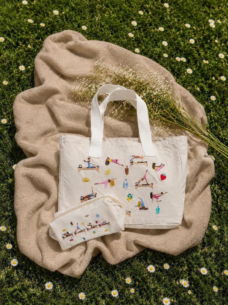 May include: A cream-colored tote bag and matching pouch set with white handles, embroidered with colorful Pilates exercise figures. The set rests on a tan blanket atop a bed of green grass with small white flowers.