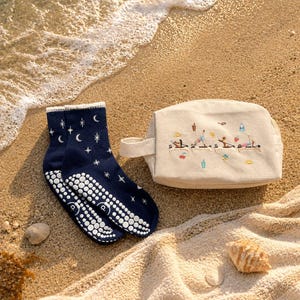 May include: Navy blue yoga socks with white grip dots and a celestial design of stars and moons. A beige canvas cosmetic bag with a handle and embroidered yoga figures is next to the socks. The items are on a sandy beach with a towel and seashells.