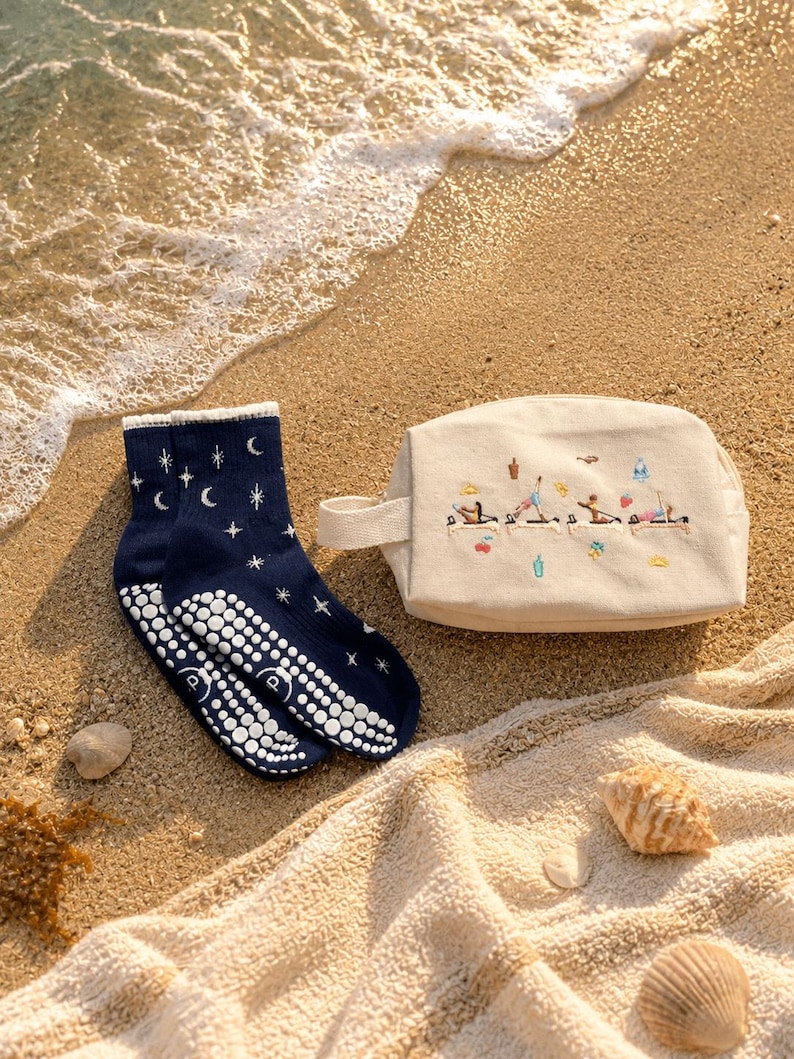 May include: Navy blue yoga socks with white grip dots and a celestial design of stars and moons. A beige canvas cosmetic bag with a handle and embroidered yoga figures is next to the socks. The items are on a sandy beach with a towel and seashells.