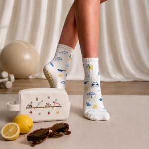 May include: White socks with blue and yellow beach-themed patterns, worn by a person. A canvas cosmetic bag with embroidered figures doing yoga, sunglasses, and lemons are in the foreground. A beige exercise ball and weights are in the background.