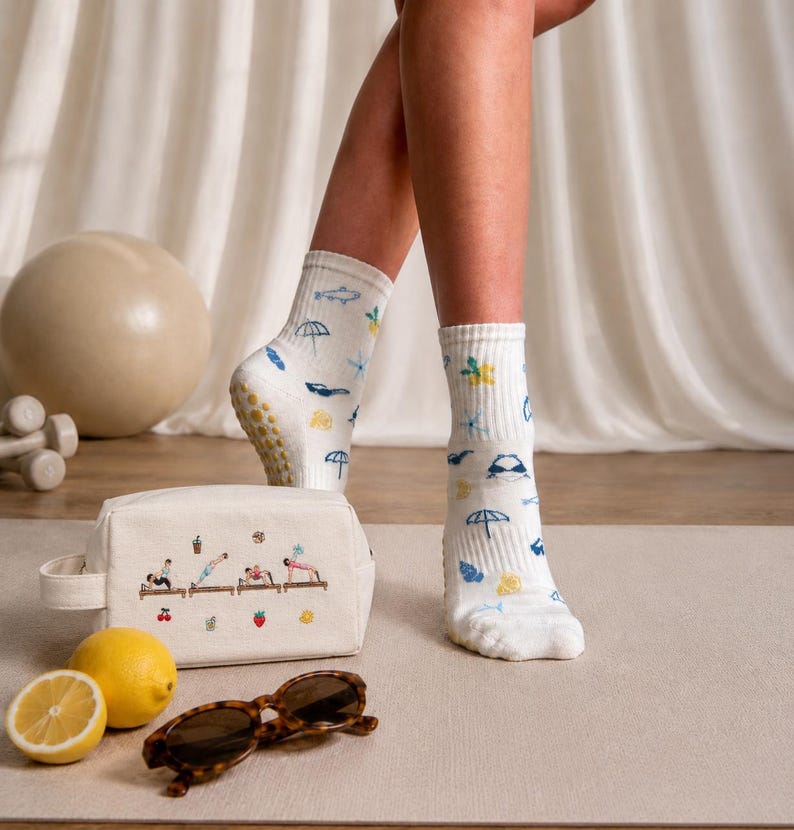 May include: White socks with blue and yellow beach-themed patterns, worn by a person. A canvas cosmetic bag with embroidered figures doing yoga, sunglasses, and lemons are in the foreground. A beige exercise ball and weights are in the background.