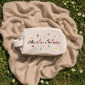 May include: A cream-colored rectangular cosmetic bag with a white handle, embroidered with colorful yoga poses. The bag rests on a beige blanket on a bed of green grass with small white flowers.