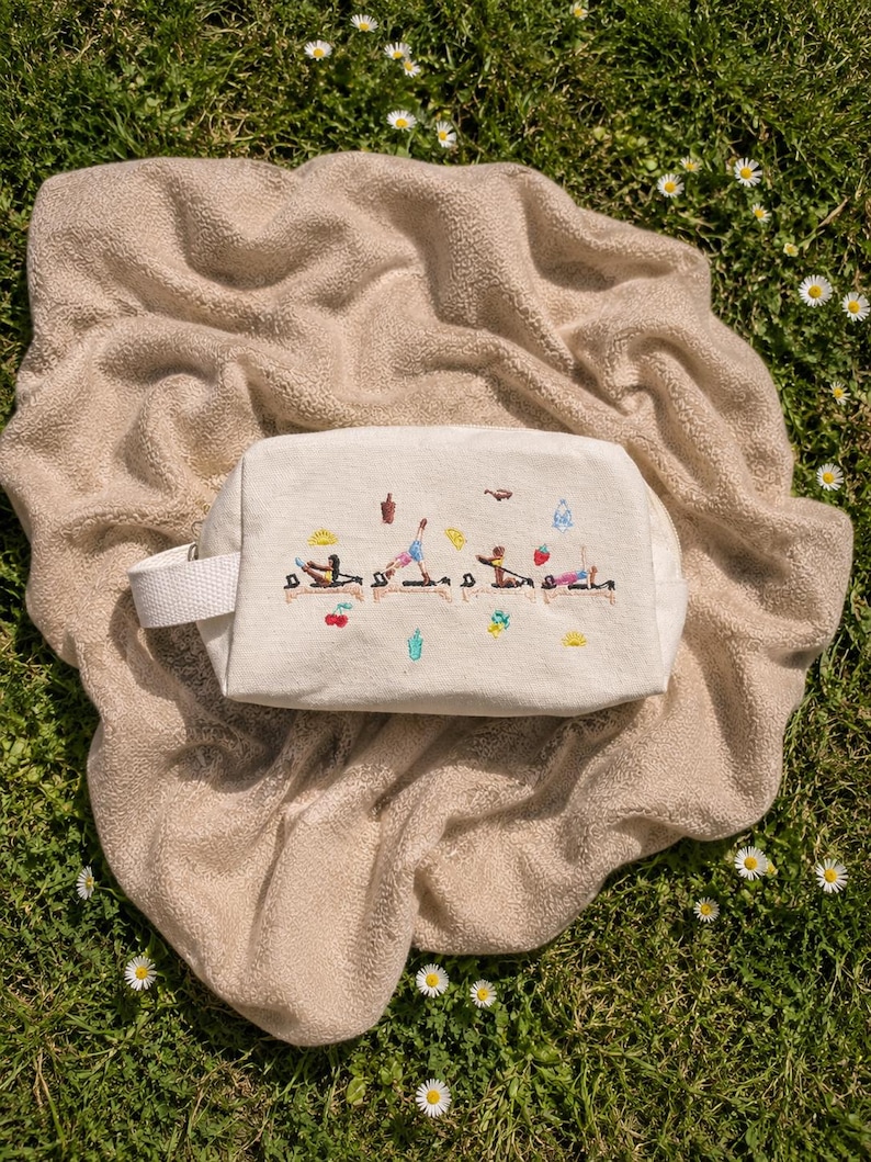May include: A cream-colored rectangular cosmetic bag with a white handle, embroidered with colorful yoga poses. The bag rests on a beige blanket on a bed of green grass with small white flowers.