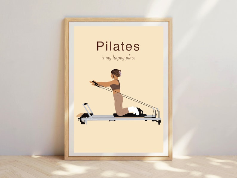 May include: Framed Pilates art print. The artwork features a woman exercising on a Pilates reformer machine. The text "Pilates is my happy place" is displayed in brown font on a cream background. The frame is a light wood color.