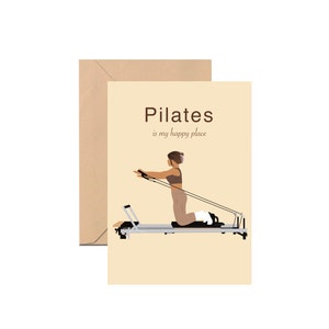 May include: A greeting card with a beige background and a Pilates theme. The card features an illustration of a person exercising on a Pilates reformer machine. The text "Pilates is my happy place" is printed on the card. A brown envelope is behind the card.