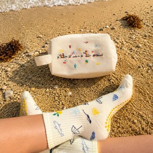 May include: A cream-colored cosmetic bag with a small handle and embroidered figures. The bag is on a sandy beach next to a pair of light yellow socks with various embroidered designs, including fish and umbrellas.