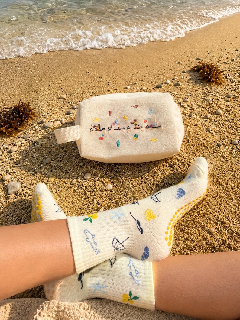 May include: A cream-colored cosmetic bag with a small handle and embroidered figures. The bag is on a sandy beach next to a pair of light yellow socks with various embroidered designs, including fish and umbrellas.