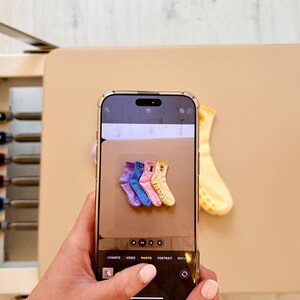 Könnte beinhalten: Ein Smartphone zeigt ein Foto von vier Paar farbigen Socken: lila, blau, rosa und gelb. Jede Socke hat ein einzigartiges Design. Eine einzelne gelbe Socke mit Muster ist ebenfalls sichtbar.