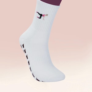 Könnte beinhalten: Weiße Sportsocken mit einer schwarzen und rosa Grafik einer Person in einer Yoga-Pose. Die Socken haben ein geripptes Design und schwarze Griffe an der Sohle. Die Socken sind für Fitnessaktivitäten konzipiert.