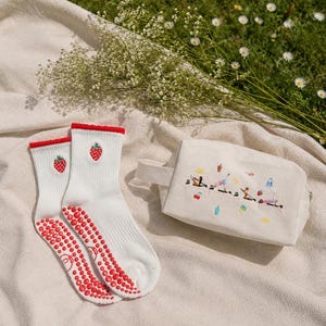 May include: White socks with red strawberry embroidery and red non-slip grips, paired with a small canvas bag featuring embroidered figures. The items are arranged on a beige blanket with a backdrop of grass and flowers.