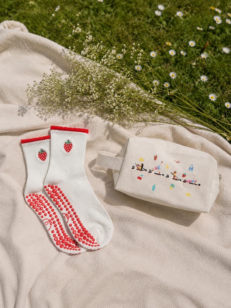 May include: White socks with red strawberry embroidery and red non-slip grips, paired with a small canvas bag featuring embroidered figures. The items are arranged on a beige blanket with a backdrop of grass and flowers.