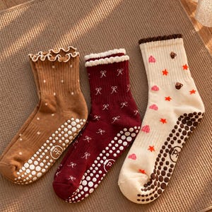 Könnte beinhalten: Drei Paar Knöchelsocken mit gerüschten Bündchen und rutschfesten Noppen an den Sohlen. Die erste ist braun mit weißen Punkten, die zweite weinrot mit weißen Schleifenmustern und die dritte cremefarben mit kleinen roten Sternen.