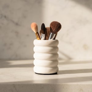 May include: A white, ribbed ceramic makeup brush holder filled with several makeup brushes. The brushes have various colors, including brown, black, and peach. The holder sits on a white surface, with a marble background.