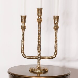 Pu&ograve; includere: Un candelabro in metallo dorato con tre portacandele. Il candelabro ha una finitura testurizzata martellata e una base larga.