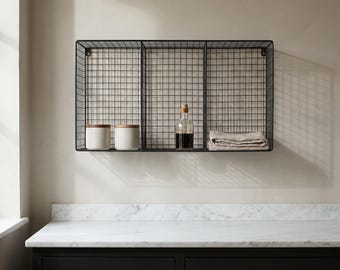 May include: A black wire mesh wall shelf with three compartments. The shelf holds two white ceramic jars with wooden lids, a clear glass bottle with a cork stopper, and a folded beige cloth. The shelf is mounted on a light-colored wall.