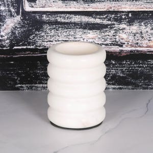 Puede incluir: Un jarrón blanco y cilíndrico con un diseño apilado y redondeado. El jarrón tiene un acabado liso y brillante y una amplia abertura en la parte superior. La base es de color oscuro. El fondo presenta un panel de madera oscura texturizada.