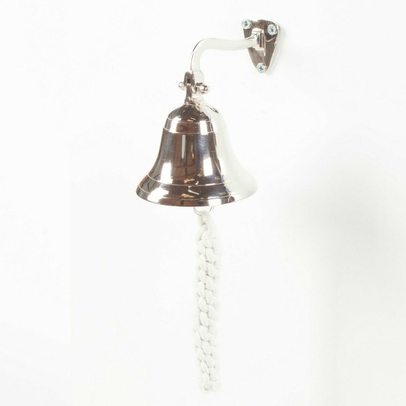 May include: A polished silver bell with a white rope pull, mounted on a wall. The bell is attached to a curved bracket and a small, triangular mounting plate with screws. The bell is a nautical-themed item.