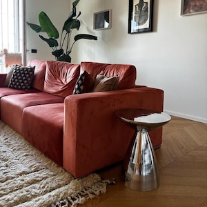 Può includere: Un divano in velluto color ruggine con profilo basso e braccioli quadrati, dotato di diversi cuscini decorativi in toni scuri e neutri, posizionato accanto a un tavolino in metallo argentato lucido con base conica e piano circolare.