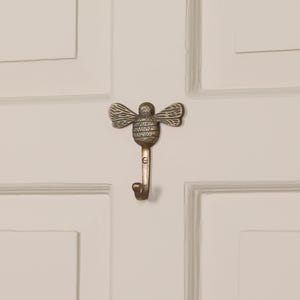Könnte beinhalten: Ein dekorativer Messing-Bienenhaken. Der Haken ist wie eine Biene mit detaillierten Flügeln und Körper geformt. Der Haken ist an einer weißen Tür befestigt. Der Haken ist ca. 10 cm lang.