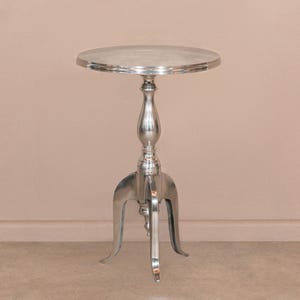 Op de afbeelding: Een zilveren metalen bijzettafel met een rond blad en drie poten. De tafel heeft een klassiek ontwerp met een gegroefde basis en een decoratief uiteinde.