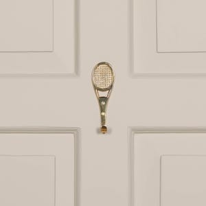May include: A gold-colored tennis racket-shaped hook. The hook has a detailed net pattern on the racket head and a curved handle with three small decorative accents. The hook is mounted on a light-colored door.