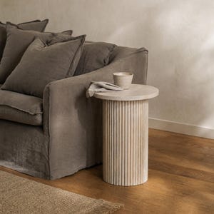 May include: A light beige side table with a round top and a cylindrical base. The base has vertical, ridged detailing. A small, light-coloured ceramic cup and a folded cloth sit on top of the table. The table is next to a grey sofa.