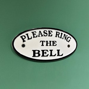 Op de afbeelding: Een wit ovaal bord met een zwarte rand en de tekst "Please Ring The Bell" op een groene achtergrond.