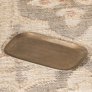 May include: A rectangular brass tray with rounded corners and a textured surface featuring a fine, cross-hatched pattern. The metal has a muted, brushed finish with a warm golden tone, and the edges are slightly raised to form a shallow basin.