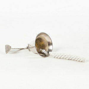 May include: A silver-colored oil lamp with a white wick. The lamp has a bell-shaped top and a small, flat handle. The wick is made of a thick, braided cotton material.