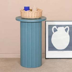 Puede incluir: Una mesa pedestal azul acanalada con una tapa redonda. La mesa tiene una cesta tejida con tres velas en la parte superior. Las velas son azules y beige. Una impresión enmarcada con el texto "Le Navire" está en el lado derecho de la mesa.