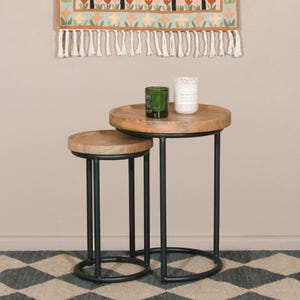 May include: A set of two nesting side tables. They have round wooden tops and black metal legs. A green glass candle holder and a white candle sit on the larger table. The background features a floral wall hanging.
