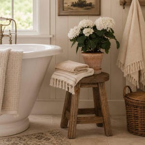 May include: A rustic wooden stool holds a terracotta pot with white hydrangea flowers, a folded beige waffle-weave towel with fringe, and a matching towel draped over a white bathtub. A woven wicker basket sits nearby in a bright bathroom.