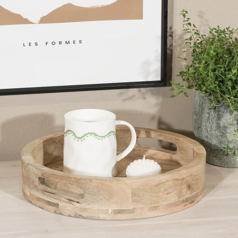 May include: A round wooden tray with a handle on the side. The tray is made of light brown wood and has a natural finish. A white mug with a green design is sitting on the tray, along with a white candle.
