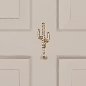 Peut inclure: Un crochet en métal doré en forme de cactus, monté sur une porte blanche. Le crochet est conçu avec des détails de cactus et une base incurvée pour suspendre des objets. C'est un objet décoratif et fonctionnel.