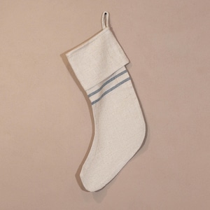 May include: A cream-coloured Christmas stocking with a folded-over cuff. Two thin, horizontal blue stripes adorn the cuff. The stocking has a loop for hanging. The fabric appears to be linen or a similar woven material.
