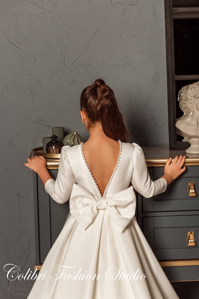 Classic ivory dress for a flower girl. The dress is made of satin. The top is decorated with pearls along the neckline and has satin sleeves. The skirt is satin and floor length. The back of the dress is complemented by a satin bow.