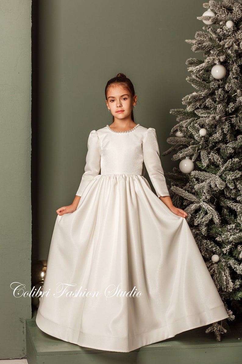 Classic ivory dress for a flower girl. The dress is made of satin. The top is decorated with pearls along the neckline and has satin sleeves. The skirt is satin and floor length. The back of the dress is complemented by a satin bow.
