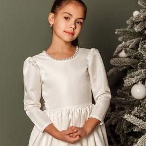 Classic ivory dress for a flower girl. The dress is made of satin. The top is decorated with pearls along the neckline and has satin sleeves. The skirt is satin and floor length. The back of the dress is complemented by a satin bow.