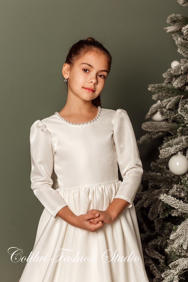 Classic ivory dress for a flower girl. The dress is made of satin. The top is decorated with pearls along the neckline and has satin sleeves. The skirt is satin and floor length. The back of the dress is complemented by a satin bow.